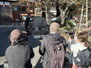 翔んでびわ湖の「大人のエコスクール」、今度は外へ出ています。ガイドの本多さんが、参加者の前で石碑の説明中です。