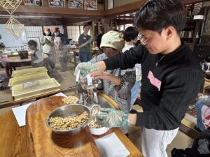 食養サークル　朝晴れ　活動のようす画像　海外からの若者と姉さんかぶりに割烹着の高齢の日本人が、協力してゆでた大豆をミンサーでつぶす作業をしている